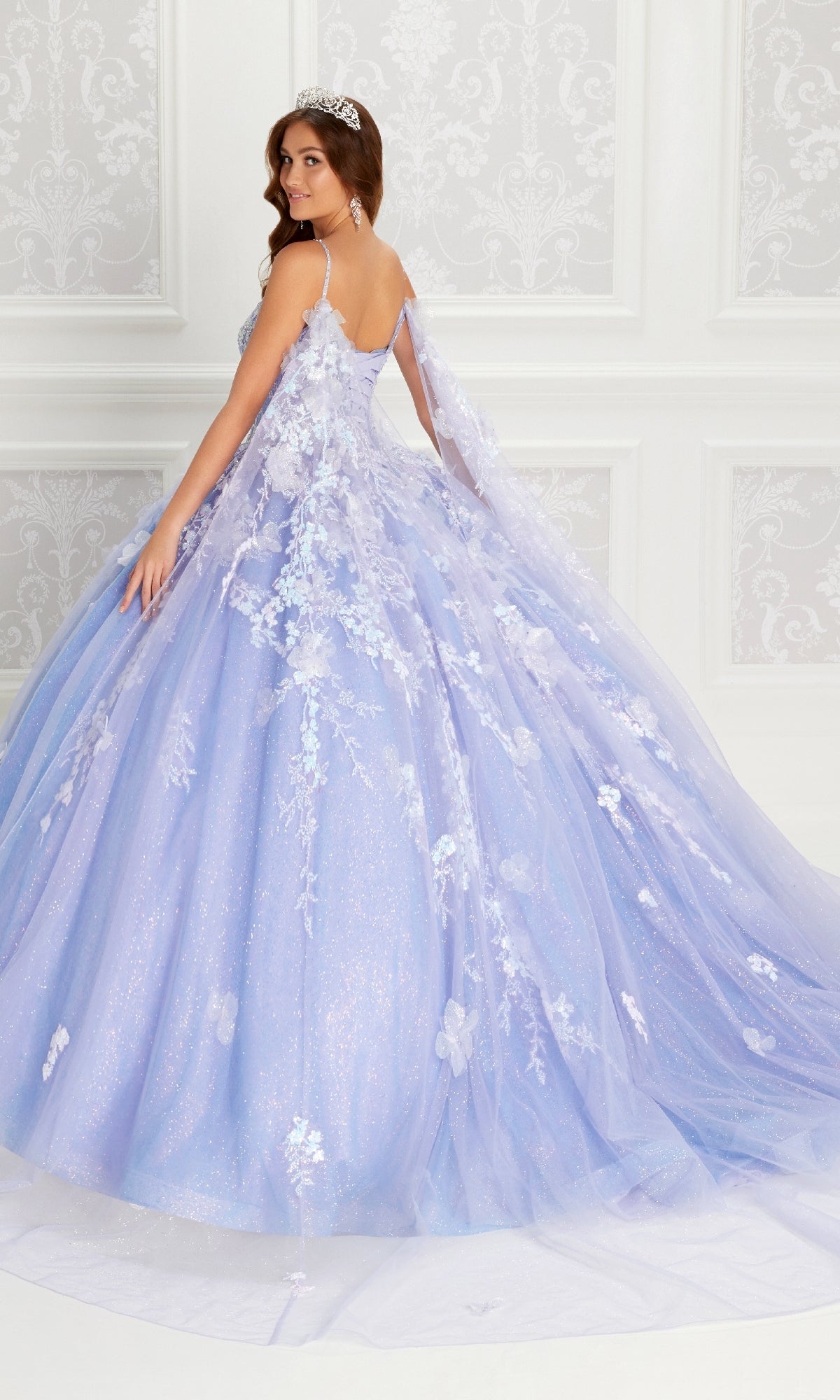 A young woman in the PR22143 By Princesa quinceanera dress, featuring sparkling lavender fabric, floral appliqués, and a beaded corset bodice, stands in an elegant white room wearing a tiara and looking over her shoulder.